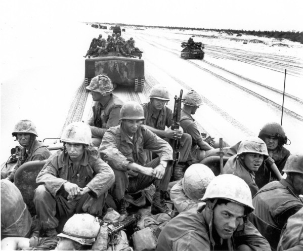 Photographic print of helmeted Marines riding atop LVTP-5 amphibious tractors moving in convoy along a sandy road, with additional vehicles visible ahead