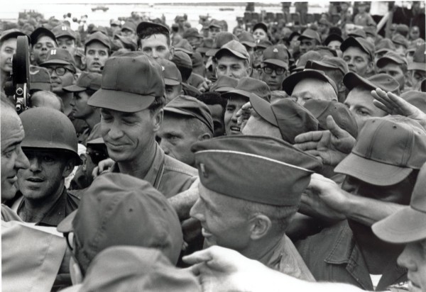 Photographic print of President Lyndon B. Johnson surrounded by a dense crowd of uniformed American troops in military caps. A film camera is visible at left as soldiers reach toward him.