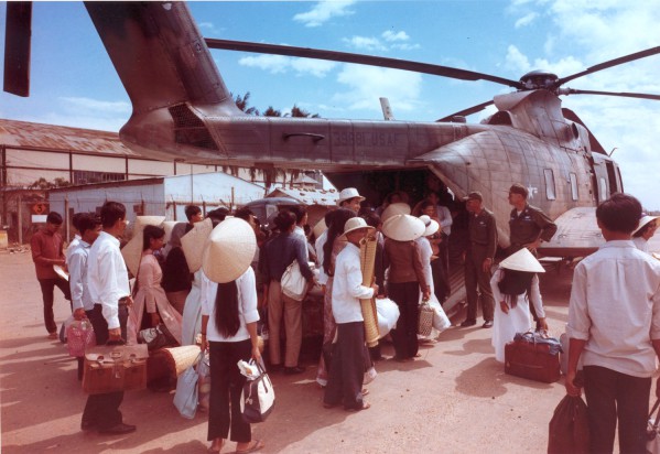Photographic print of a crowd of villagers carrying bags, baskets, and luggage boarding a large USAF CH-3C helicopter on a dusty airfield
