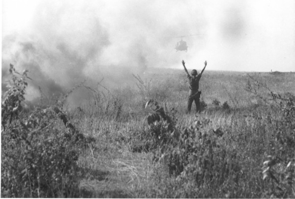 Photographic print of a helmeted U.S. Army soldier standing in a grassy field with both arms raised, signaling a UH-1 helicopter approaching through billowing smoke