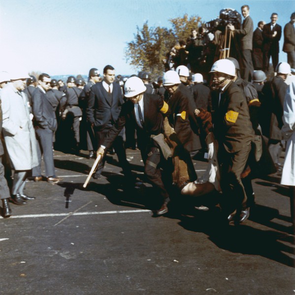 Photographic print of U.S. Marshals in white helmets and yellow armbands dragging a limp protester across pavement, surrounded by a crowd of onlookers and press