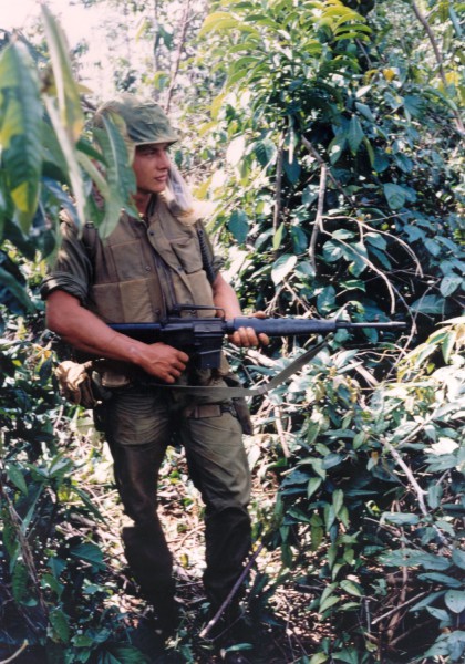 Photographic print of a soldier in olive fatigues and flak vest holding a rifle with fixed bayonet, standing amid dense tropical foliage