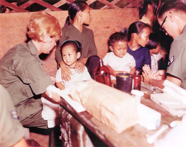 Photographic print of a uniformed woman examining a young child seated on a table indoors, with medical supplies and other children and adults nearby