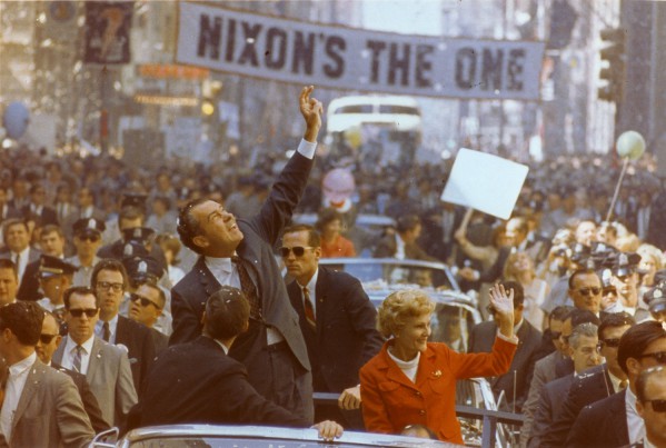 Photographic print of Richard and Pat Nixon standing in an open convertible, waving to crowds beneath a banner reading "Nixon's The One"
