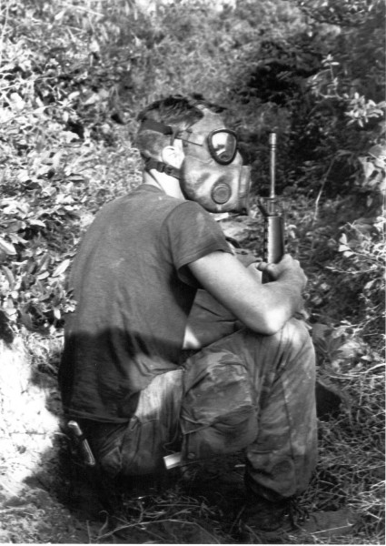 Photographic print of a soldier in camouflage pants and a gas mask crouching outdoors, holding a rifle, surrounded by dense foliage