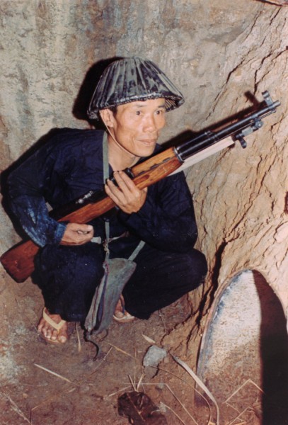 Photographic print of a soldier crouching in an earthen bunker, wearing dark clothing and a patterned hat, holding a wooden-stocked SKS rifle