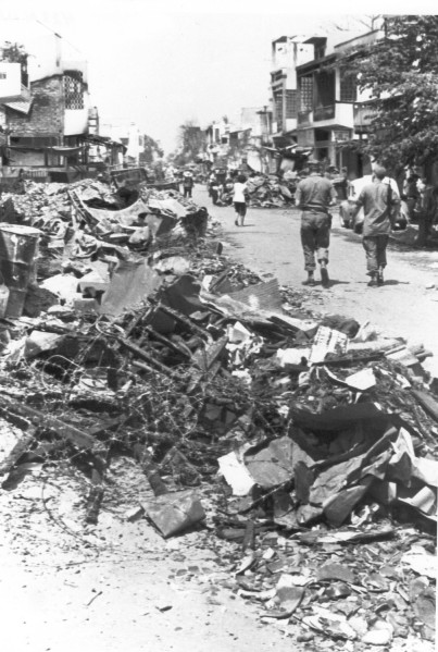 Photographic print of a street in Cholon, Saigon, lined with large piles of rubble and tangled barbed wire. Partially damaged buildings stand in the background as several figures walk down the debris-strewn road