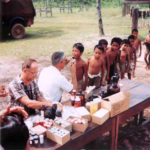 Photographic print of two men at an outdoor table covered with medicine bottles and boxes examining a line of young shirtless children in a rural setting