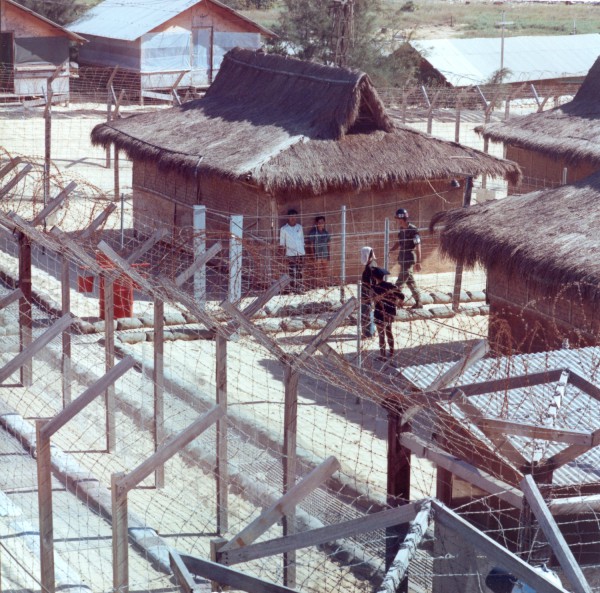 Photographic print of a fenced compound with thatched-roof structures and extensive barbed wire. A helmeted military policeman walks among several detainees near a sandbag barrier