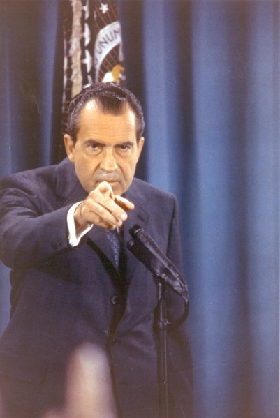 Photographic print of President Nixon in a dark suit pointing toward the camera at a microphone stand, with a blue curtain and presidential flag behind him