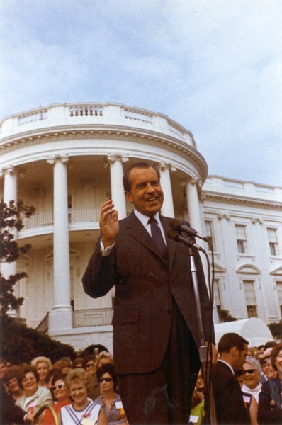 Photographic print of President Richard Nixon speaking at microphones on the White House south lawn, smiling with one hand raised, a crowd gathered behind him
