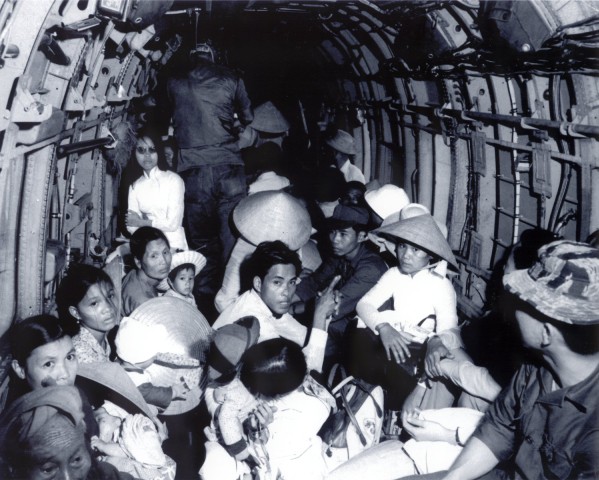 Photographic print of a large group of Vietnamese refugees, including children, crowded inside a military helicopter cargo bay. Many wear conical straw hats and carry bundles