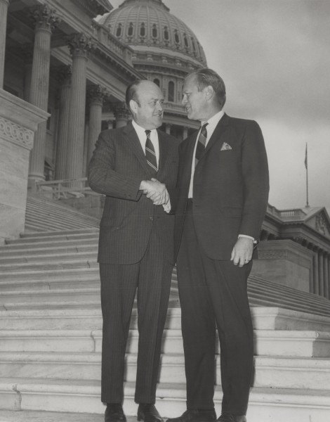 Photographic print of two men in suits shaking hands on the steps of the U.S. Capitol building, its dome visible behind them