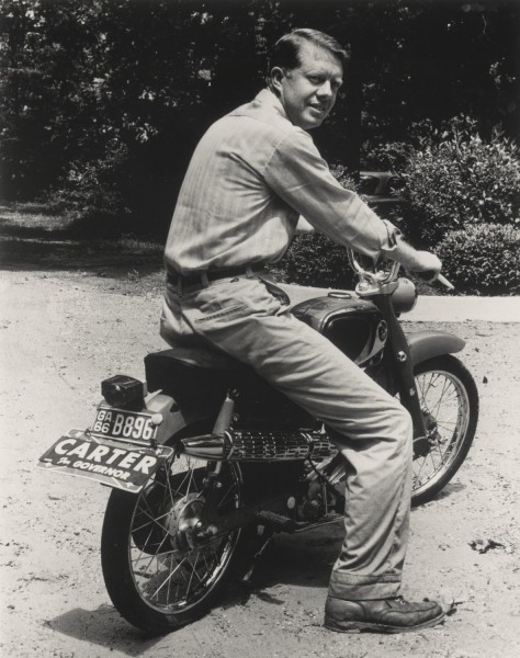 Photographic print of Jimmy Carter seated on a motorcycle with a "Carter for Governor" campaign plate attached below a Georgia 1966 license plate