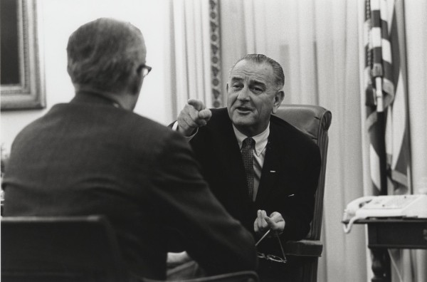 Photographic print of LBJ seated in a leather chair, pointing toward an unidentified man whose back faces the camera, an American flag visible at right