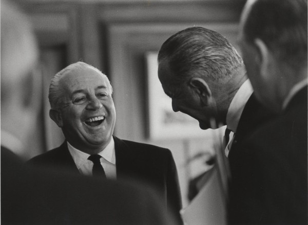 Photographic print of two men in dark suits sharing a laugh, one facing the camera with a wide smile, the other leaning in from the right