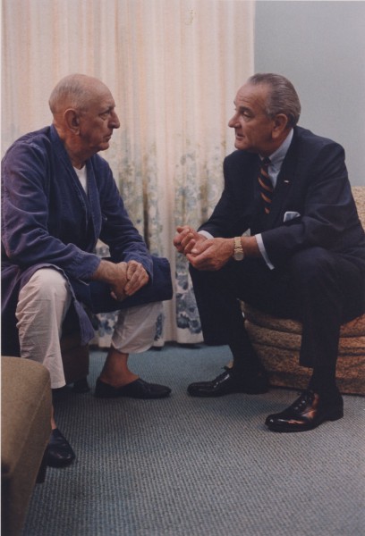 Photographic print of two men seated facing each other in conversation. One wears a blue robe and pajamas, the other a dark suit with a striped tie