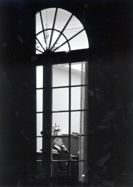 Photographic print of President Lyndon Johnson seated at a desk holding a telephone, shot through a tall multi-pane window with a fanlight above