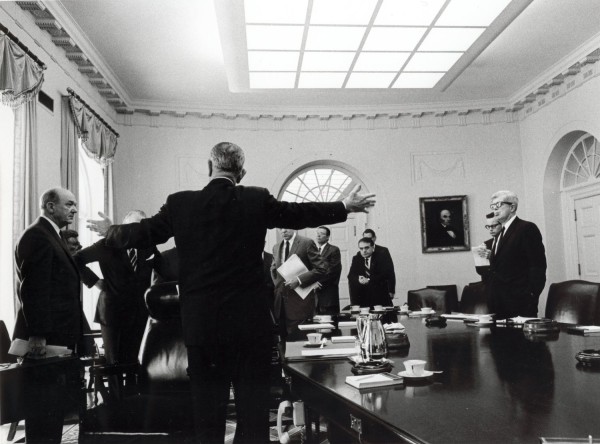 Photographic print of President Lyndon Johnson, back to camera and arms outstretched, addressing a group of suited men gathered around a large conference table in the Cabinet Room