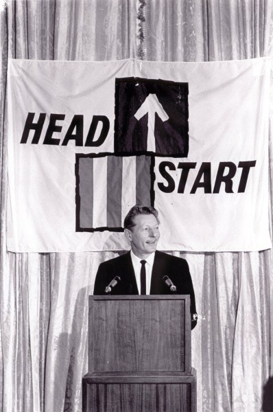 Photographic print of Danny Kaye speaking at a wooden podium with two microphones, a large "Head Start" banner hanging behind him on draped curtains