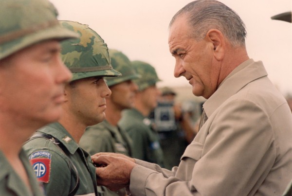 Photographic print of President Lyndon B. Johnson pinning a medal on a young soldier wearing an 82nd Airborne patch, with a line of helmeted troops behind them