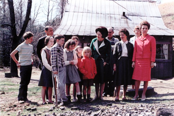 Photographic print of a group of adults and children gathered outdoors in front of a rustic wooden building with a metal roof on a sunny day
