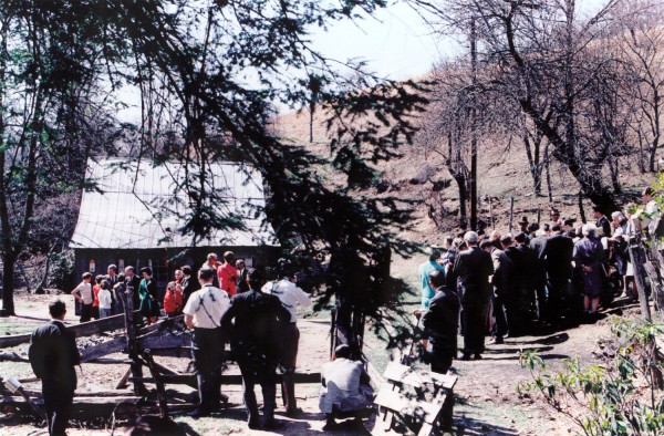 Photographic print of a large crowd gathered outdoors near a small rural cabin surrounded by bare trees on a hillside