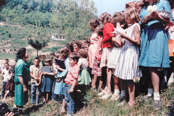 Photographic print of Lady Bird Johnson in a teal dress addressing a large group of schoolchildren outdoors on a grassy hillside with trees behind
