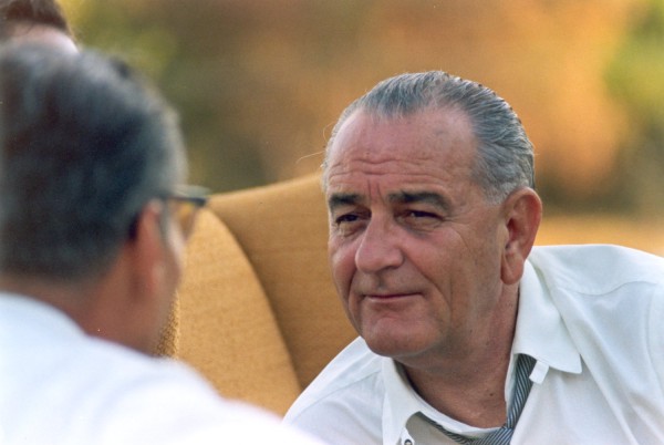 Photographic print of President Lyndon B. Johnson wearing a white shirt with unbuttoned collar and loosened tie, seated outdoors with a blurred background