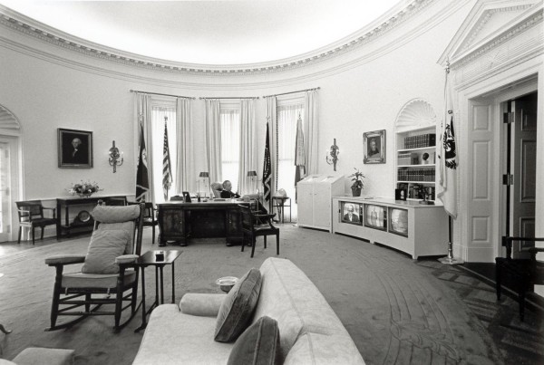 Photographic print of the Oval Office with a man seated at the presidential desk facing a bank of three televisions along the wall