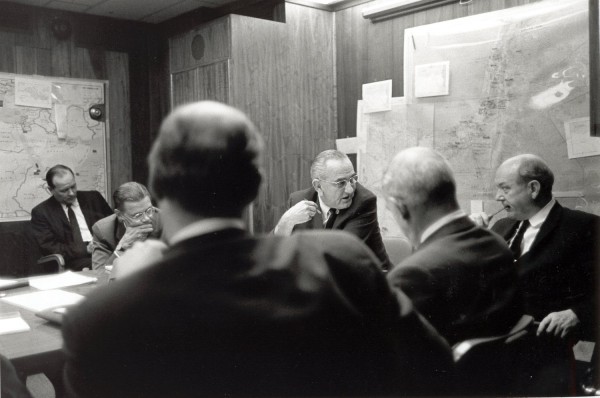 Photographic print of several men in suits seated around a conference table in a wood-paneled room, with large maps mounted on the wall behind them