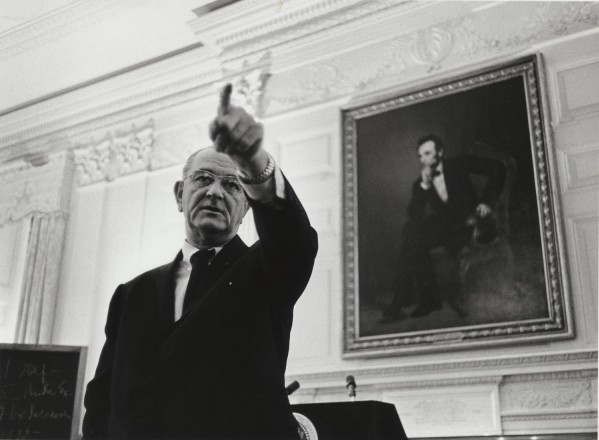 Photographic print of President Lyndon Johnson pointing upward in a formal room. A framed portrait hangs on the white ornate wall behind him.