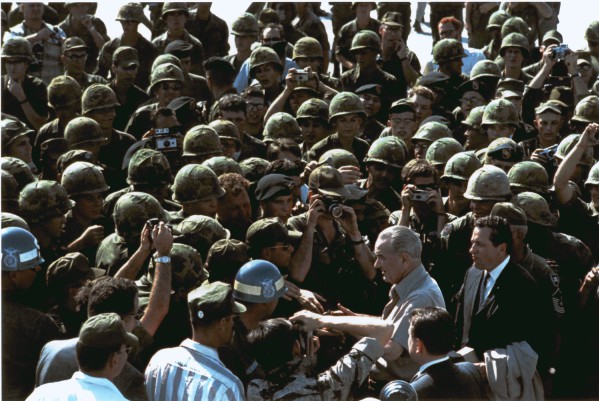 Photographic print of President Lyndon B. Johnson shaking hands amid a dense crowd of soldiers in helmets and fatigues, many holding cameras