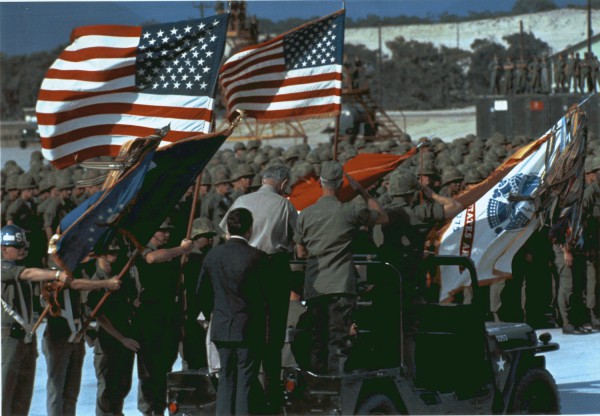 Photographic print of a large formation of helmeted American troops at a military base, with multiple American and unit flags waving. Civilians and officers stand near a military jeep in the foreground