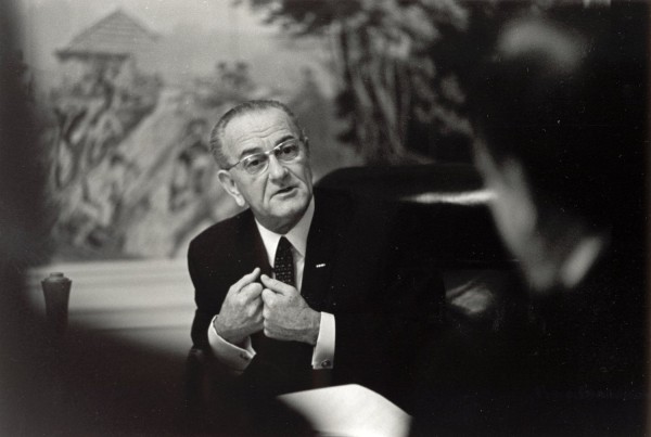 Photographic print of President Lyndon B. Johnson in a dark suit and glasses, gesturing while speaking across a table to another person