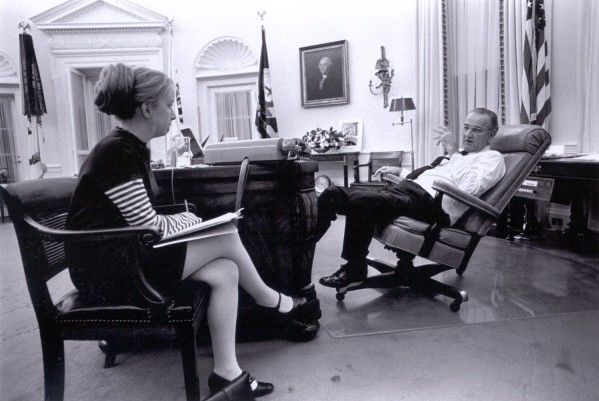 Photographic print of two people seated facing each other in a formal office, one holding a notepad, the other reclined in a leather chair near a large desk and framed portrait