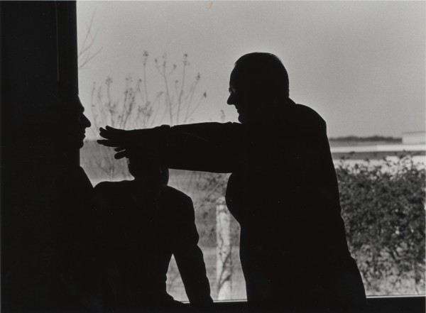Photographic print of LBJ in dark silhouette reaching one arm outward toward another silhouetted figure, with a window and bare trees behind