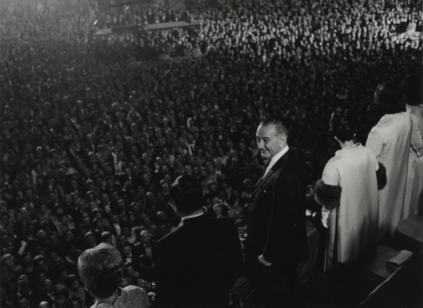 Photographic print of a smiling man in a dark suit on a stage, facing a massive indoor crowd filling the frame. Several formally dressed figures stand nearby on the right.