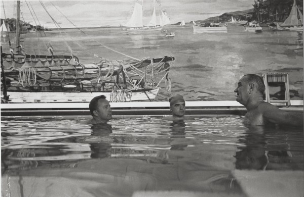 Photographic print of three people submerged in an indoor pool, conversing. A large nautical mural with sailboats covers the wall behind them