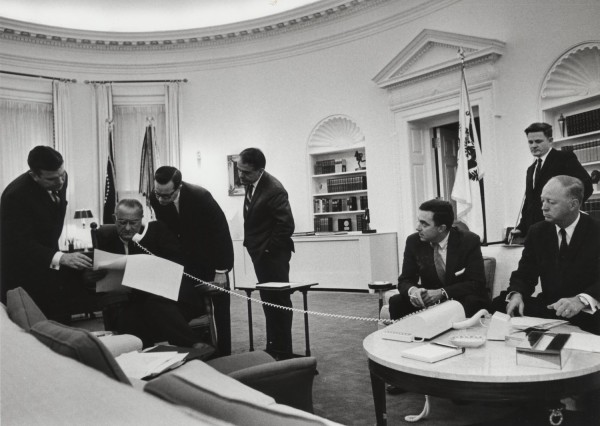 Photographic print of LBJ seated in the Oval Office, speaking on the phone while advisers lean in to review documents around him