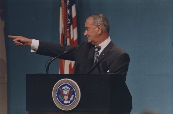 Photographic print of Lyndon B. Johnson in a dark suit at a presidential podium, arm extended and pointing to the left, with an American flag behind him
