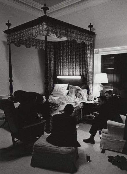 Photographic print of a man reclining in a canopied bed, surrounded by several seated figures in a dimly lit bedroom