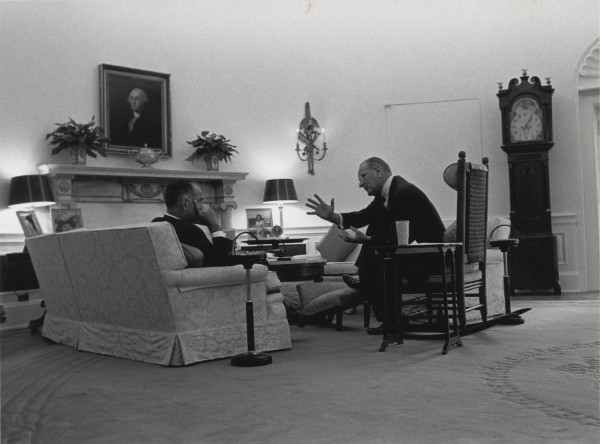 Photographic print of two suited men seated in the Oval Office, one gesturing expressively. A grandfather clock, portrait painting, and fireplace are visible in the background