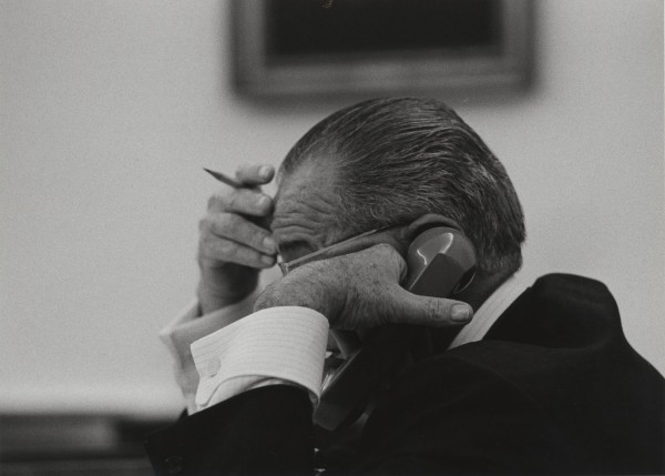 Photographic print of a man in a dark suit holding a telephone receiver to his ear, a pencil raised to his forehead, framed artwork visible on the wall behind