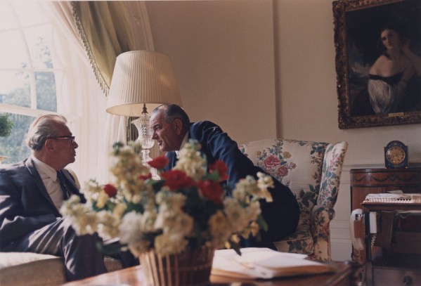 Photographic print of LBJ leaning forward in a floral armchair to speak with Sen. Dirksen in an ornate room with a painting and flower arrangement