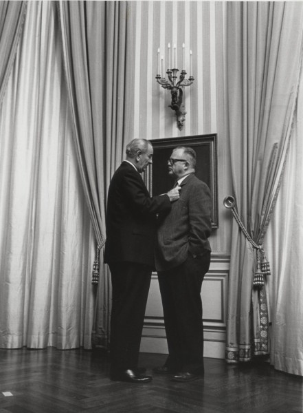 Photographic print of two men in suits standing in a formal room with draped curtains, one grasping the other's lapel while facing him