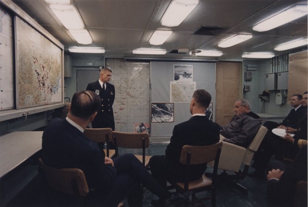 Photographic print of several men seated in a briefing room while a uniformed naval officer stands before large wall maps and aerial photographs