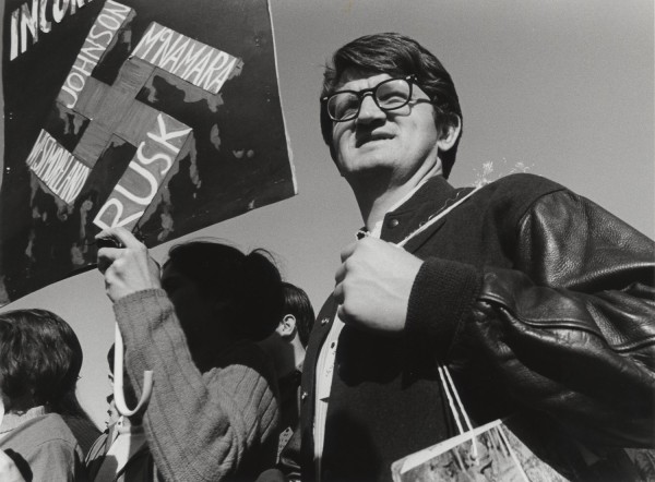 Photographic print of a young man wearing glasses and a varsity jacket standing beside a protest poster bearing a swastika with the names Johnson, McNamara, Rusk, and Westmoreland