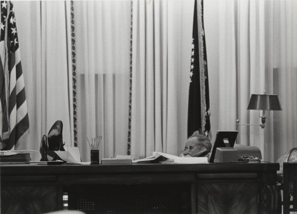 Photographic print of Lyndon B. Johnson talking on the phone at his desk, feet propped up, with American flags and curtained windows behind him
