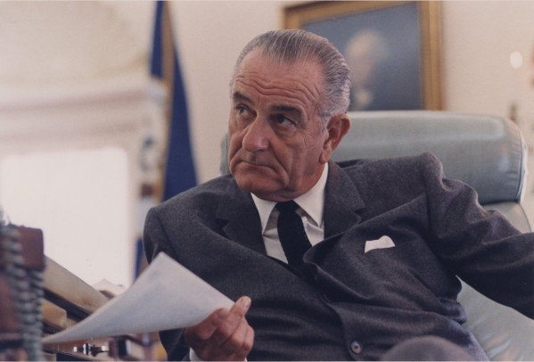 Photographic print of Lyndon B. Johnson seated in a leather chair, holding a document, wearing a dark suit and tie. A blue flag and framed painting are visible in the background